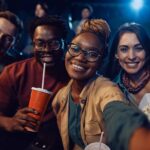 Happy African American woman and her friends taking selfie during movie premiere in a theater and looking at camera.